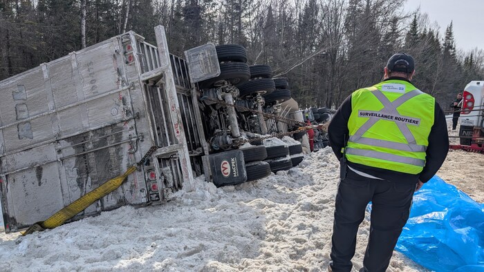 Un homme de dos portant un dossard de surveillance routière est debout près d’un camion renversé aux abords d’une autoroute et d’un boisé. 