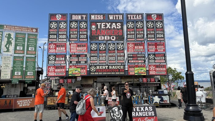 Un kiosque nommé Texas Rangers BBQ exhibe de nombreux prix devant plusieurs personnes rassemblées à l'extérieur.