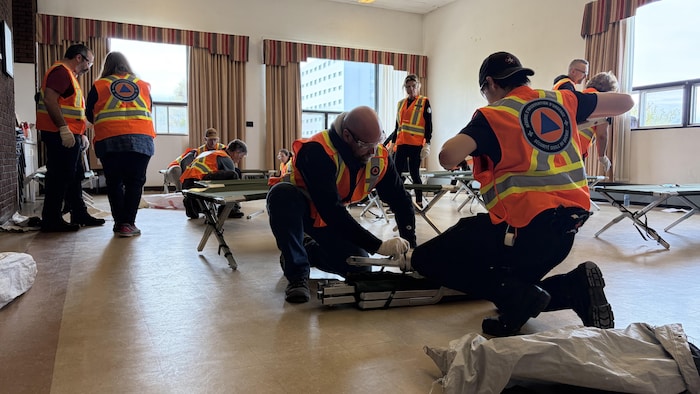 Plusieurs personnes portant des dossards avec le logo de la Sécurité civile au dos sont réunies deux par deux, dans un atelier de formation. Elles travaillent à monter des lits de camps dans une salle.