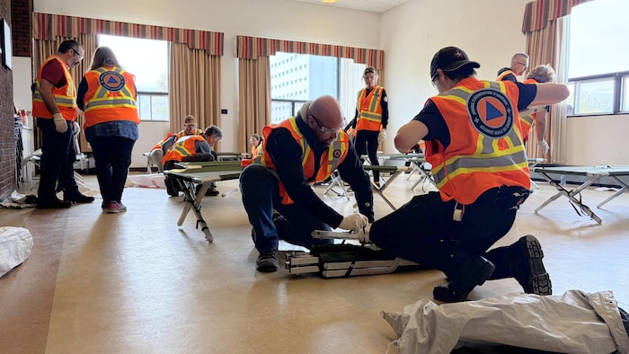 Plusieurs personnes portant des dossards avec le logo de la Sécurité civile au dos sont réunies deux par deux, dans un atelier de formation. Elles travaillent à monter des lits de camp dans une salle.  
