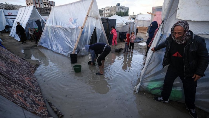 Dans un campement de tentes de fortune, des hommes s'affairent à remplir des seaux d'eau tandis que des enfants les observent.