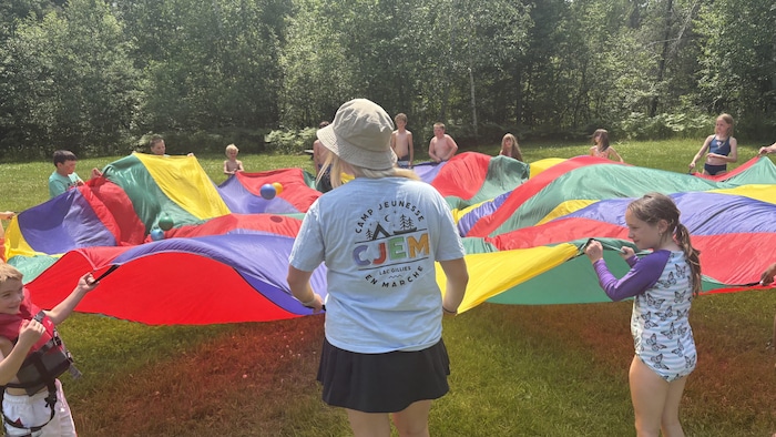 Une monitrice et son groupe de jeunes campeurs jouent avec des ballons et un parachute multicolore.