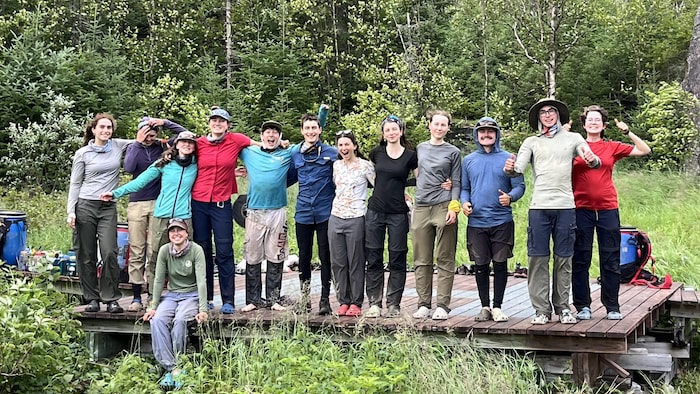 Une douzaine d'ados et de jeunes adultes sourient à la caméra, tous joyeux. Ils sont vêtus de vêtements de plein air pour faire une expédition de canot camping. Il se trouve sur une plateforme de bois à proximité d'une forêt.