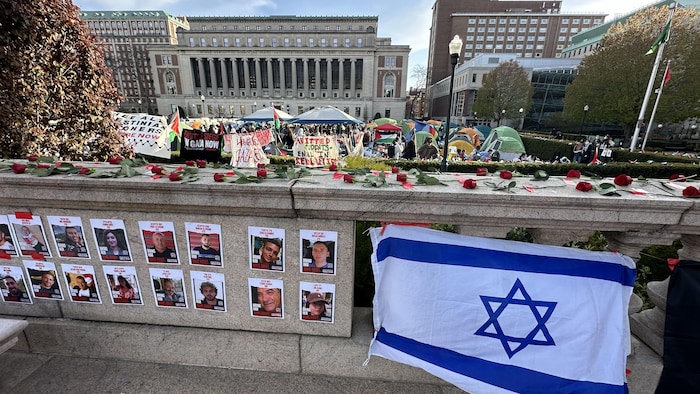 Le campement propalestinien sur le terrain de l'Université Columbia, avec des tentes, des drapeaux palestiniens et des banderoles, derrière un muret sur lequel se trouvent les photos des otages, un drapeau d'Israël et des roses.