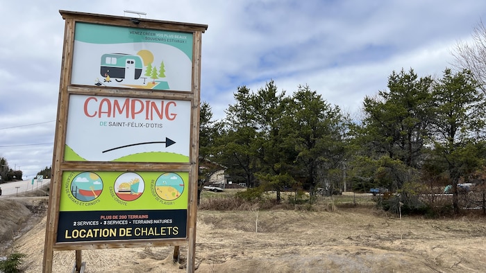 Une grande enseigne en bois annonce le Camping de Saint-Félix-d'Otis. Le panneau présente des illustrations colorées, mentionne la location de chalets et de kayaks, ainsi qu'un casse-croûte, devant un paysage de forêt et un ciel nuageux.