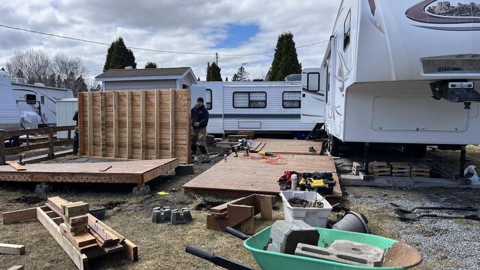 Un homme travaille à la déconstruction d'une terrasse en bois sur un terrain de camping. Des outils, une brouette et des matériaux jonchent le sol entre plusieurs roulottes garées sous un ciel nuageux.