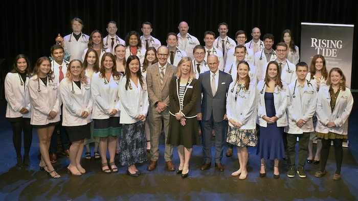 Une photo de groupe de 30 jeunes femmes et jeunes hommes en sarrau blanc, stéthoscopes autour du cou, disposés sur trois rangées et entourant une dirigeante et deux