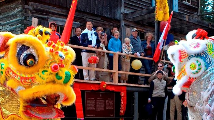 People are watching an art show in front of a wooden house. We also see two flags raised: the Canadian and the Chinese.