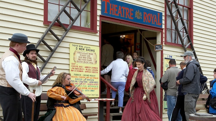 A man plays a banjo, a woman plays a fiddle and another woman dances in front of the entrance to a theater as tourists enter the building. The musicians are dressed in traditional 19th century clothing.
