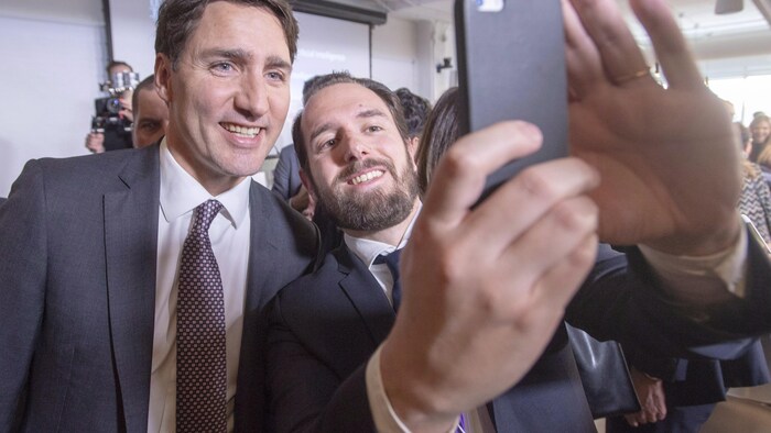 Le premier ministre Justin Trudeau posant pour un égoportrait avec un jeune homme barbu. Les deux hommes portent un veston-cravate.