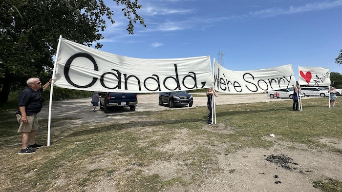 Quatre personnes tenant trois bannières où l’on peut lire « Canada, nous sommes désolés. Michigan » dans un stationnement au pied du pont Gordie-Howe à Détroit, le 24 juillet 2026, lors de l’inauguration du pont.