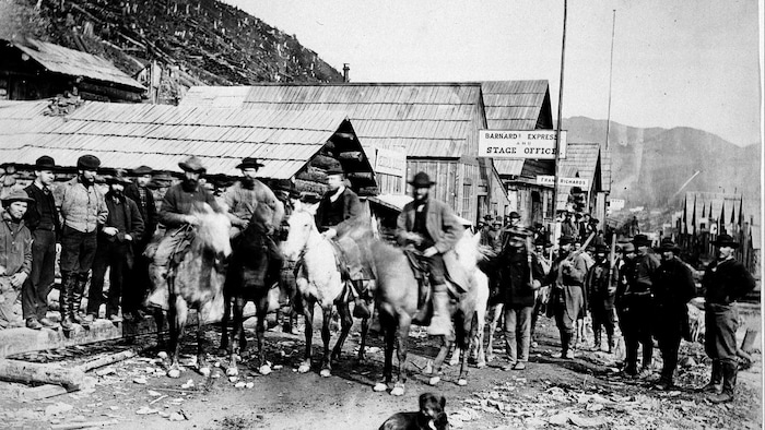 Photo en noir et blanc d'un groupe d'hommes rassemblés sur la rue principale de Barkerville. Certains sont debout et d'autres sont à cheval.