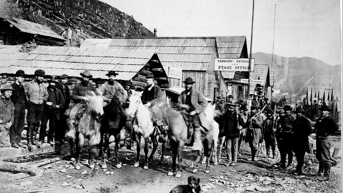 Black and white photo of a group of men gathered on Barkerville's main street. Some are standing and some are on horseback.