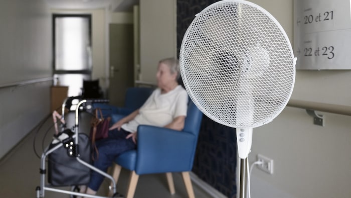 Un ventilateur sur pied au premier plan dans un couloir où une femme âgée est assise à côté d'un déambulateur.