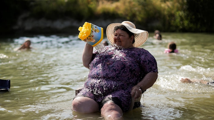 Une femme en robe mauve, coiffée d'un grand chapeau de paille, est assise dans une rivière. Suant comme une champlure, elle fait une grimace en versant sur sa poitrine l'eau contenue dans un seau jaune pour enfants.
