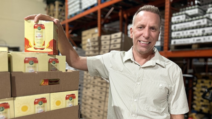 Un homme souriant en chemise à manches courtes pose dans un entrepôt, s'appuyant sur une pile de boîtes de bière de la microbrasserie « La Voie Maltée », de la variété Lager Mexicaine.