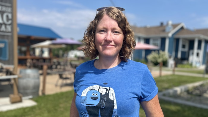 Portrait extérieur montrant une femme souriante aux cheveux roux bouclés. Elle porte un t-shirt bleu avec une illustration graphique. L'arrière-plan flou révèle la terrasse d'une microbrasserie avec des parasols et des bâtiments sous un ciel ensoleillé.