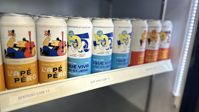 Plusieurs canettes de bière de la Microbrasserie du Lac-St-Jean, notamment les variétés « L'Apéro » et « ¡Que Viva Lac-St-Jean! », sont alignées sur l'étagère d'un réfrigérateur de magasin avec des étiquettes de prix de 5,50$.
