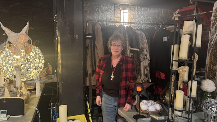 Une femme se tient debout devant un rack de costumes. À droite et à gauche, des décorations lumineuses qui serviront pour le festival du Canoë volant.
