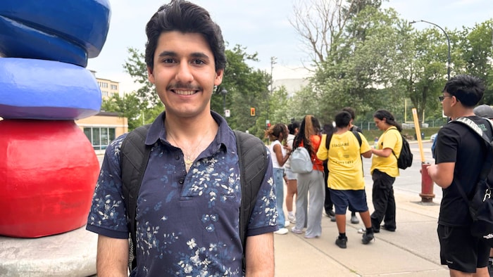 Un jeune homme souriant pose devant le CHU Sainte-Justine, à Montréal, devant un groupe d'élèves du secondaire.