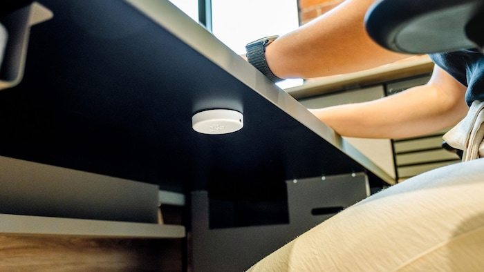 Cette vue en contre-plongée montre un petit capteur circulaire blanc fixé sous le plateau d'un bureau noir. Une personne portant une montre intelligente est assise devant l'espace de travail dans un environnement de bureau moderne avec un mur en briques.