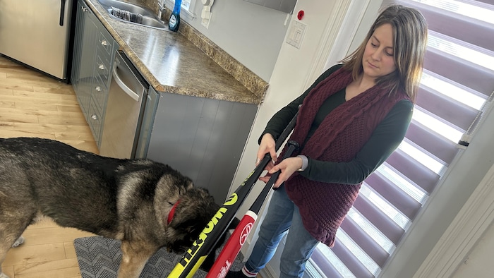 Une femme tient deux bâtons de baseball, devant une porte, son chien à côté d'elle.