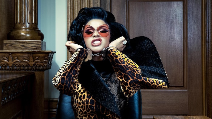 Cardi B porte une tenue léopard, de grandes lunettes aux verres roses et ses mains sont menottées. 