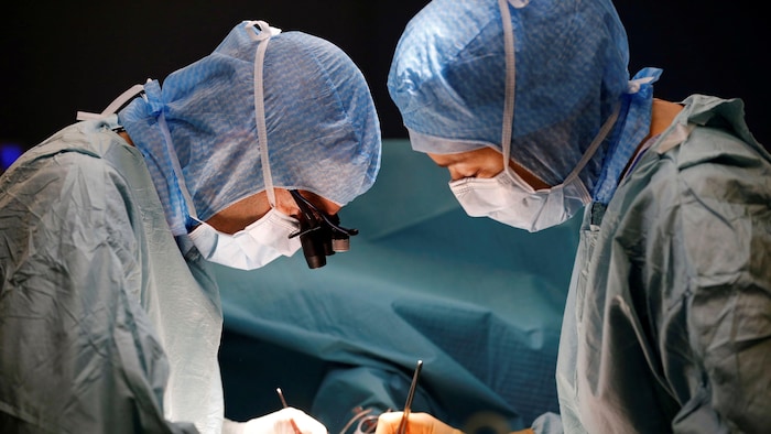 Dans une salle d'opération, deux personnes portant des combinaisons bleues et des masques chirurgicaux sont face à face, penchées au-dessus d'une table d'opération où est couché un patient, hors du cadre de la photo.