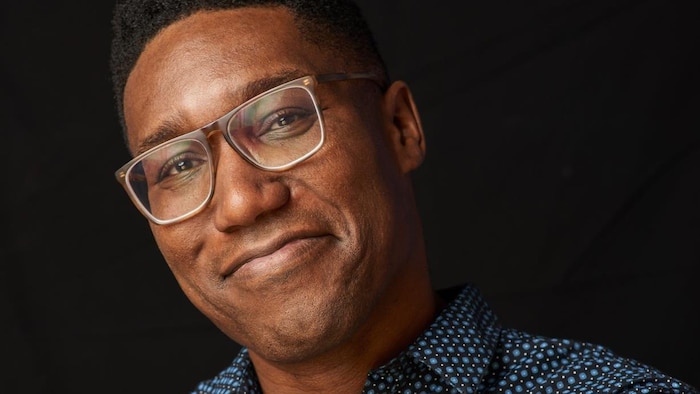 Portrait d'un homme souriant avec des lunettes et une chemise bleue sur un fond noir.