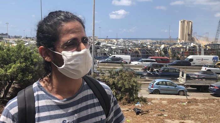 Un homme portant un masque et des lunettes se tient près d'une route. Loin derrière lui, on aperçoit les silos endommagés par les explosions.