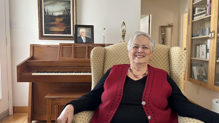Carmelle Lebreux assise dans un fauteuil. Derrière elle, le portrait de Marcel Cormier est installé sur un piano.