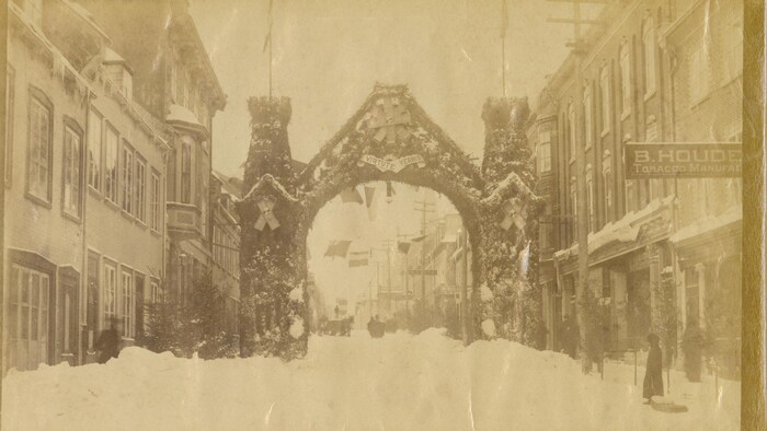 Une arche se dresse sur toute la largeur de la rue Saint-Jean, couverte de neige après une tempête. On aperçoit plusieurs passants et des banderoles suspendues entre les commerces.