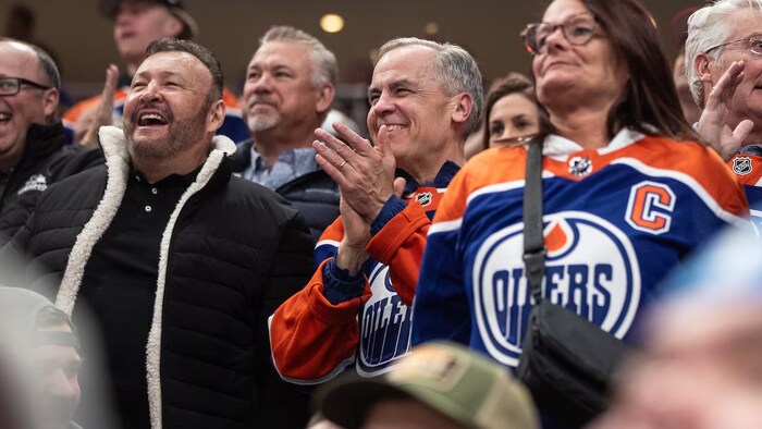 Mark Carney dans une foule de partisans sportifs. Une femme près de lui est vêtue d'un maillot des Oilers.