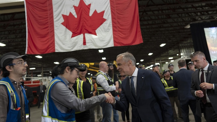 Mark Carney serre la main d'un ouvrier. Un drapeau canadien est accroché en arrière-plan.