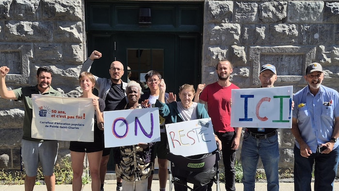 Des membres du Carrefour d'éducation populaire de Pointe-Saint-Charles brandissent des pancartes indiquant « On reste ici! ».