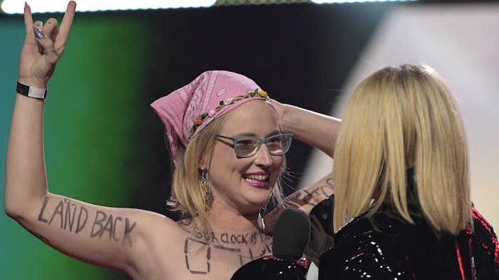Une femme portant un foulard rose sur la tête regarde une femme blonde de dos, avec un air de défi. 