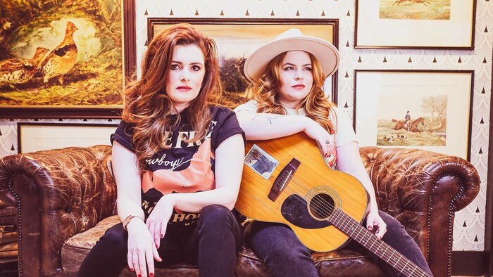 Deux soeurs assises sur un sofa à deux places, dans une pièce au mur décoré de plusieurs illustrations représentant des animaux. La femme assise à gauche regarde droit dans la caméra, et celle de droite, qui porte un chapeau de cow-boy et est appuyée sur une guitare sèche, regarde de côté.