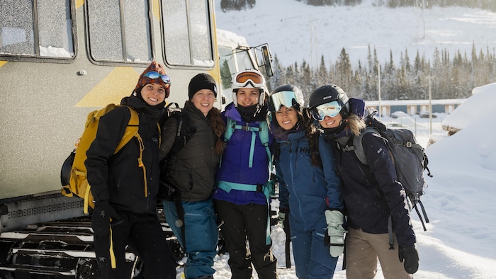 Un groupe de femme en combinaison de ski souris à l'objectif, devant une dameuse.