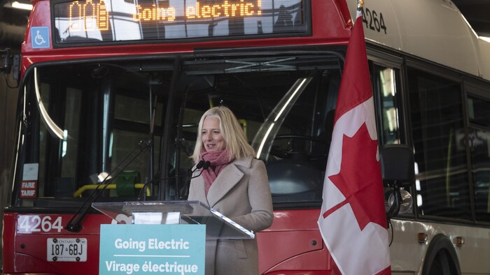 Catherine McKenna devant un autobus et un drapeau canadien. Elle parle devant un lutrin sur lequel une pancarte dit : Virage électrique.