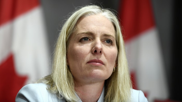 Une femme aux cheveux mi-longs regarde droit devant elle, l'air calme. Derrière elle sont accrochés des drapeaux du Canada.