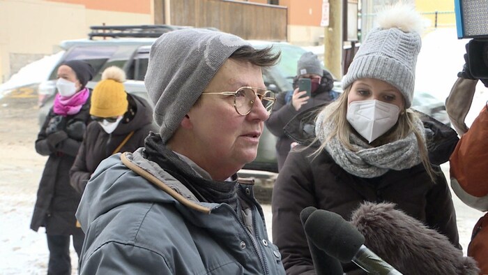 Une femme avec une tuque et un manteau est interviewée par des journalistes dehors dans le centre-ville.