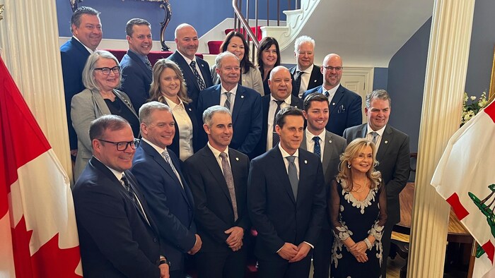 Dix-huit personnes sont debout sur trois rangées pour une photo de groupe au bas d'un escalier. À gauche sur la photo, on voit un cellulaire tenu à l'horizontale par quelqu'un qui prend une photo.