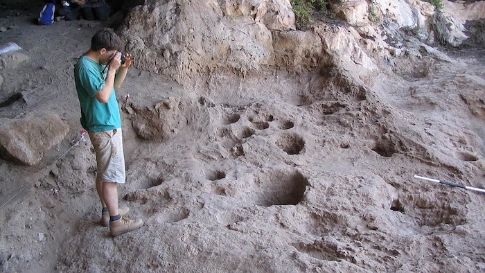 On voit les cavités creusées dans le sol de la caverne de Raqefet. À gauche, un homme prend une photo.