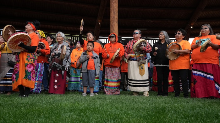 Des membres de la Première Nation Tk'emlúps te Secwépemc vêtus de chandails orange jouent du tambour lors de la cérémonie marquant le premier anniversaire de la découverte de sépultures anonymes près de l'ancien pensionnat pour Autochtones de Kamloops, en Colombie-Britannique, le lundi 23 mai 2022.
