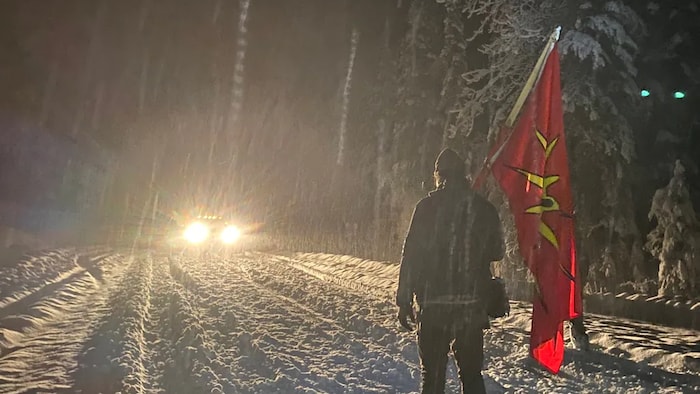 Un manifestant tenant un drapeau des guerriers mohawks devant une voiture aux phares allumés, la nuit, sur une route des environs de Prince George, en Colombie-Britannique.