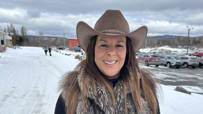 Une femme qui porte un chapeau de cowboy et un col de fourrure est debout dans la neige et sourit en regardant l'objectif de la caméra.