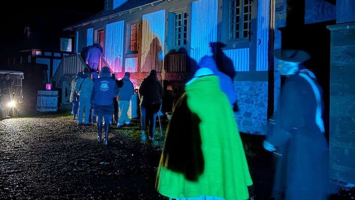Un groupe de gens vêtus de façon moderne se dirige vers un bâtiment en pierre. Ils sont suivis de deux silhouettes en costumes du 18e siècle.