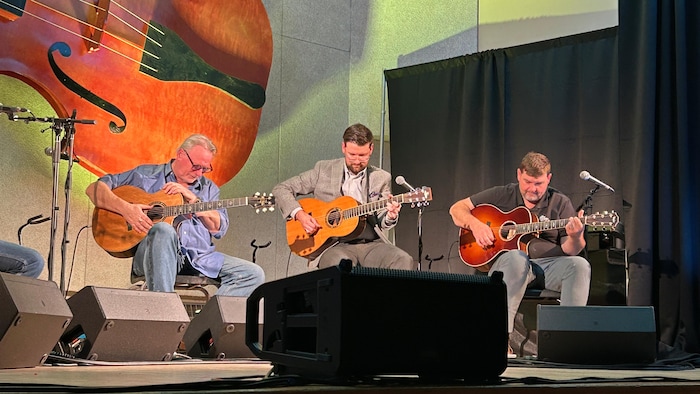 Sur une scène dont le mur est décoré d'un immense violon, trois guitaristes sont sur scène, assis. Celui à gauche incline la tête et affiche un air attentif pendant que les deux autres jouent.