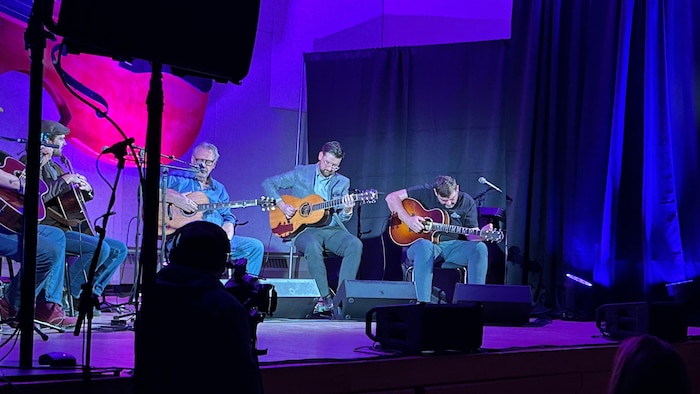 Cinq guitaristes assis sur scène avec leur instrument, jouant et s'écoutant mutuellement.