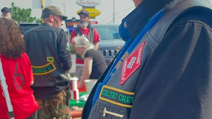 Un groupe dehors avec l'enseigne du Tim Hortons en arrière-plan. À l'avant, on voit un écusson rouge avec le chiffre 81 en blanc sur une veste de cuir.